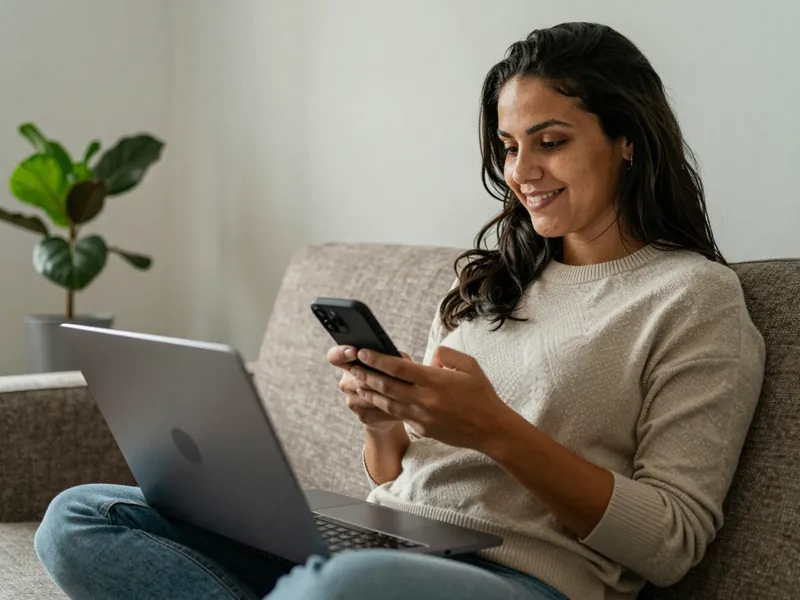 Afiliada no sofá com notebook conferindo o celular